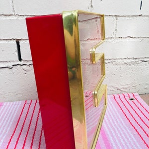 May include: A red and gold vintage sign with the letter "E" in a clear glass panel. The sign is made of metal and has a rectangular shape.