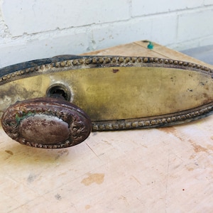May include: Antique brass door plate with a decorative border and a matching knob. The plate is oval shaped and has a keyhole. The knob is round and has a detailed design.