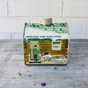 May include: A vintage maple syrup tin shaped like a cabin. The tin features a painted scene of a person standing outside a cabin door, with the words "ABSOLUTELY PURE MAPLE SYRUP" across the top. The tin holds 16.9 fl oz.