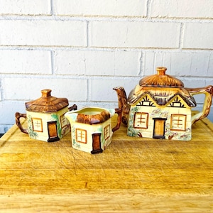 May include: A ceramic tea set featuring a teapot, creamer, and sugar bowl, each designed to resemble a quaint cottage. The set has a yellow base with brown roof details, windows, and doors. The teapot measures approximately 8 inches in height.