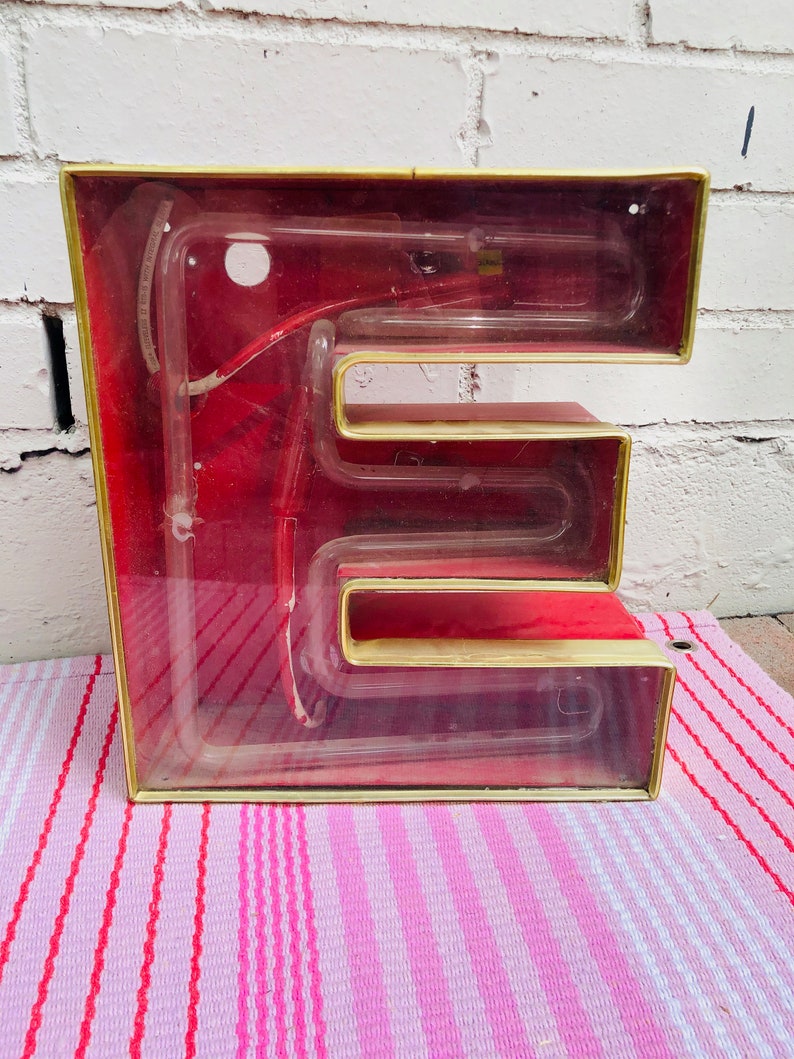 May include: A vintage neon sign in the shape of the letter "E". The sign is made of glass and has a gold frame. The neon tube is visible inside the sign, and the sign is lit up with a red glow.