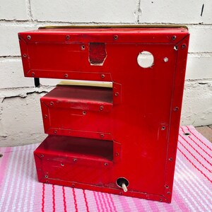 May include: Red metal letter E with a gold top. The letter is made of individual pieces of metal that are riveted together. The letter is sitting on a pink and white striped surface.