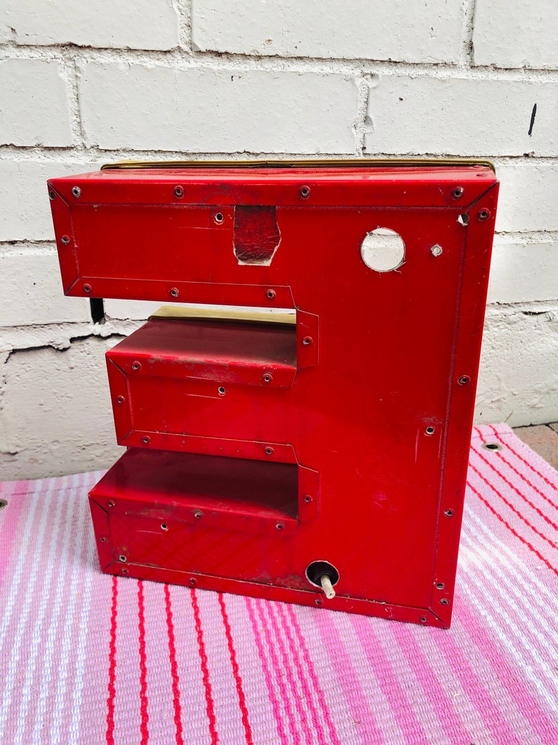 May include: Red metal letter E with a gold top. The letter is made of individual pieces of metal that are riveted together. The letter is sitting on a pink and white striped surface.