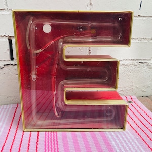May include: A vintage red and gold neon sign in the shape of the letter E. The sign is made of glass and has a clear plastic cover. The neon tubing is visible inside the sign.