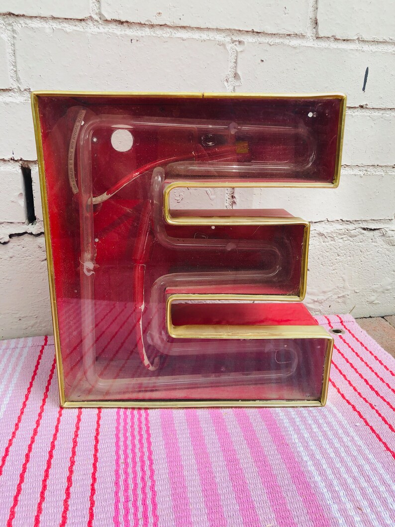 May include: A vintage red and gold neon sign in the shape of the letter E. The sign is made of glass and has a clear plastic cover. The neon tubing is visible inside the sign.