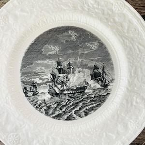 May include: White ceramic plate with a black and white nautical scene in the center. The image depicts ships at sea, possibly in battle. The plate has a decorative embossed floral pattern around the rim.