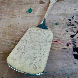 May include: A silver cake server with an ornate, engraved design. The server has a long, slender handle and a wide, flat blade.