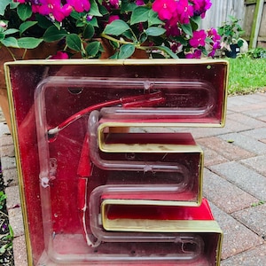 May include: A vintage neon sign in the shape of the letter "E". The sign is made of glass and metal and has a red background with gold trim. The neon tubing is visible inside the sign.