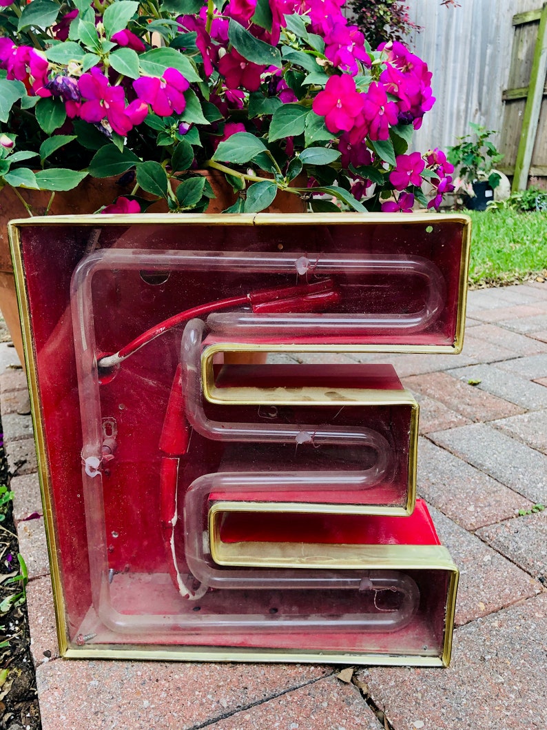 May include: A vintage neon sign in the shape of the letter "E". The sign is made of glass and metal and has a red background with gold trim. The neon tubing is visible inside the sign.
