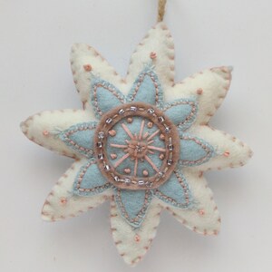 May include: A white felt star ornament with a blue and pink flower design and beaded accents.