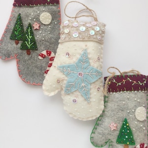 May include: Three felt Christmas ornaments shaped like mittens. The mittens are decorated with different winter themes, including a snowflake, a Christmas tree, and a candy cane.