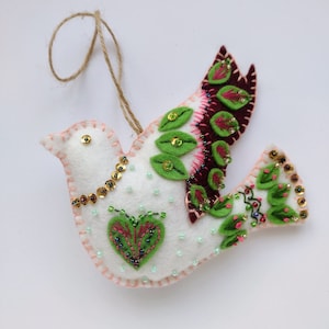 May include: White felt dove ornament with green and burgundy embroidered leaves and a heart shape. The dove is decorated with gold sequins and green beads.