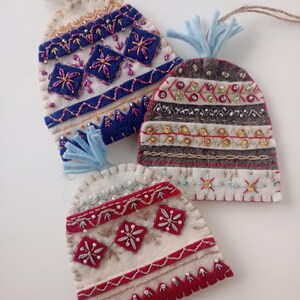May include: Three felt ornaments shaped like winter hats with tassels. One is white with blue geometric embroidery and beads, one is grey with gold sequins and silver beads, and one is white with red trim, red diamond shapes, and bead accents.