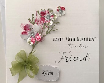 May include: A white card with a pink and white floral design and the text "Happy 70th Birthday to a dear Friend" and "Sylvia".
