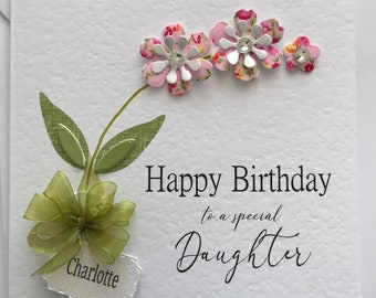 May include: A white card with a green bow and leaves, three pink flowers with silver glitter centers, and the text "Happy Birthday to a special Daughter Charlotte".