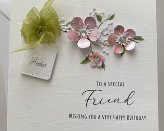 May include: A white birthday card with a textured surface, featuring pink paper flowers with crystal centers and green leaves. A green ribbon and a tag that says "Heather" are attached. The card reads "To a Special Friend, Wishing You a Very Happy Birthday."