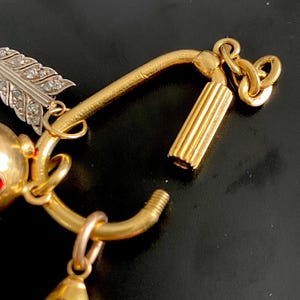 Puede incluir: Primer plano de una pulsera de dijes dorada con un colgante de pez, una pluma con detalles de diamantes y un cierre cil&iacute;ndrico. La pulsera tiene una est&eacute;tica vintage con detalles intrincados y un acabado pulido.