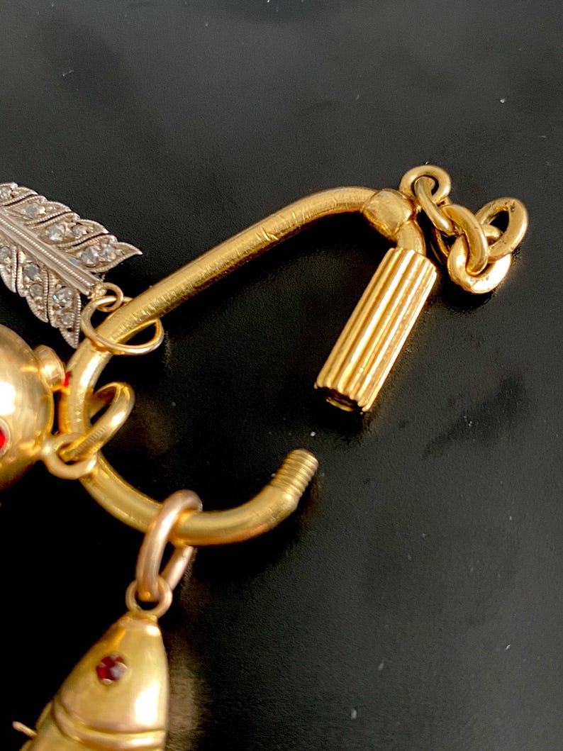 Puede incluir: Primer plano de una pulsera de dijes dorada con un colgante de pez, una pluma con detalles de diamantes y un cierre cil&iacute;ndrico. La pulsera tiene una est&eacute;tica vintage con detalles intrincados y un acabado pulido.
