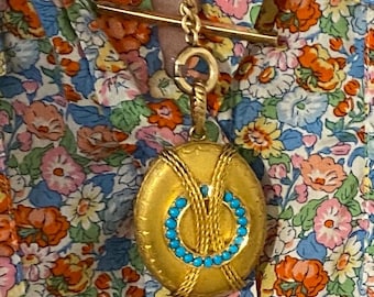 Puede incluir: Un colgante de medallón ovalado en tono dorado con un círculo central de pequeñas piedras turquesas y detalles texturizados tipo cuerda, suspendido de una cadena dorada con cierre de barra horizontal, sobre una camisa de flores.