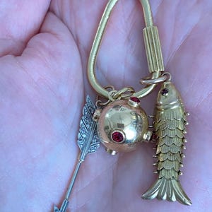 Puede incluir: Colgante dorado con un pez, una esfera con gemas rojas y una flecha plateada con dise&ntilde;o de pluma. El colgante cuelga de un lazo dorado. El pez tiene escamas detalladas y la esfera tiene tres gemas rojas.
