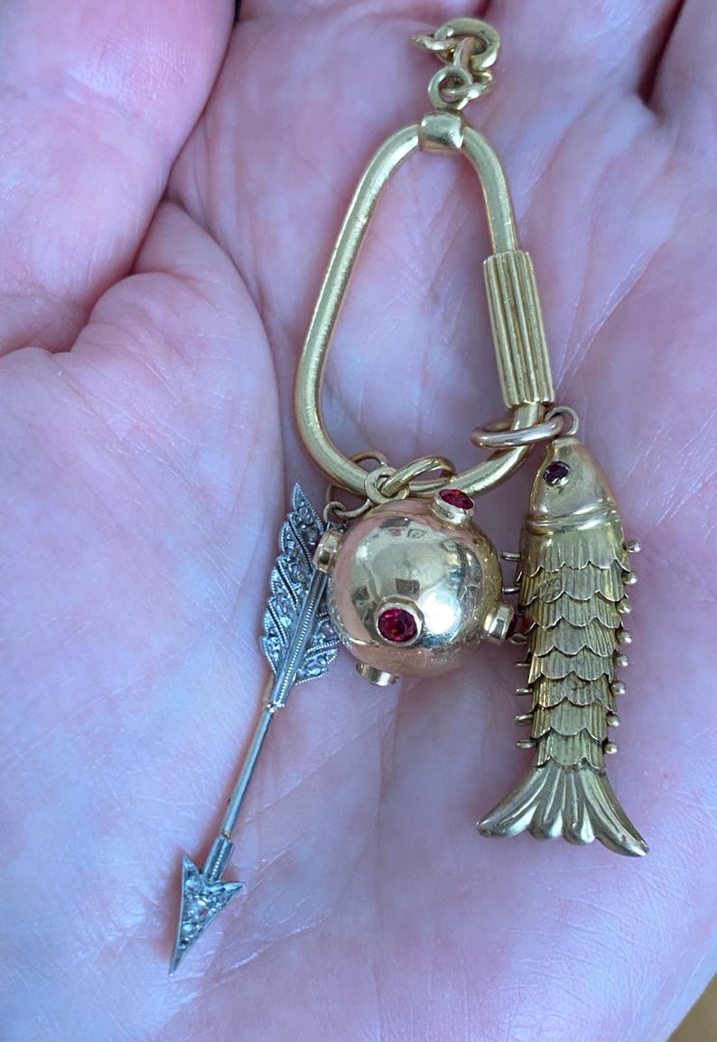 Puede incluir: Colgante dorado con un pez, una esfera con gemas rojas y una flecha plateada con dise&ntilde;o de pluma. El colgante cuelga de un lazo dorado. El pez tiene escamas detalladas y la esfera tiene tres gemas rojas.