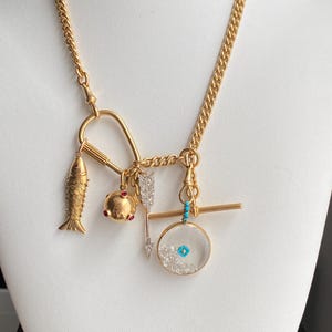 Puede incluir: Un collar dorado con m&uacute;ltiples colgantes. Los colgantes incluyen un pez, un colgante redondo con detalles rojos, una flecha y un colgante redondo transparente con detalles azules y blancos. El collar tiene una cadena de eslabones.