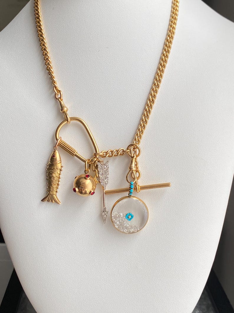 Puede incluir: Un collar dorado con m&uacute;ltiples colgantes. Los colgantes incluyen un pez, un colgante redondo con detalles rojos, una flecha y un colgante redondo transparente con detalles azules y blancos. El collar tiene una cadena de eslabones.