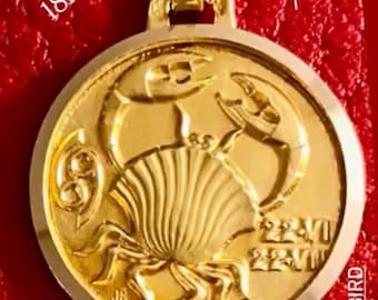 Puede incluir: Un colgante dorado del signo zodiacal Cáncer, en oro de 18 quilates. El colgante presenta un diseño detallado de un cangrejo y mide aproximadamente 15 mm de diámetro. El texto "French, Cancer zodiac charm in 18kt" es visible. También hay una pequeña bandera francesa.