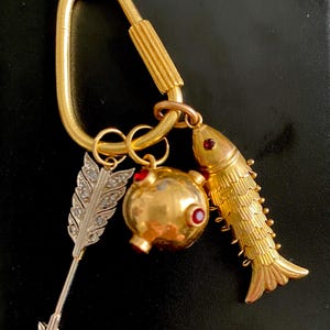 Puede incluir: Llavero dorado con colgantes decorativos. Los colgantes incluyen un pez, un objeto redondo con detalles rojos y una pluma plateada con adornos de diamantes. El llavero tiene un cierre tipo mosquet&oacute;n.