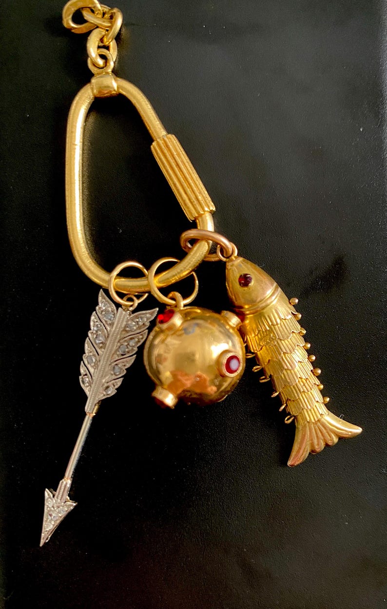 Puede incluir: Llavero dorado con colgantes decorativos. Los colgantes incluyen un pez, un objeto redondo con detalles rojos y una pluma plateada con adornos de diamantes. El llavero tiene un cierre tipo mosquet&oacute;n.