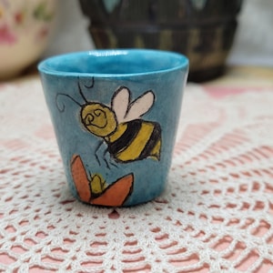 May include: A small, hand-painted ceramic cup with a turquoise exterior. The cup features a whimsical illustration of a cartoon bee with yellow and black stripes, and a flower. The cup is on a lace doily.
