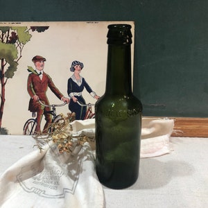 May include: A vintage green glass bottle with a label that reads "J.C.G. & Son" on a white cloth with a vintage advertisement for a bicycle company in the background.