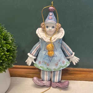 May include: A ceramic clown ornament with a blue and white outfit, featuring a ruffled collar and a matching hat. The ornament has a gold string for hanging and is decorated with floral accents and purple shoes.