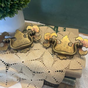 Peut inclure: Deux poignées de tiroir en laiton dotées d'une plaque arrière décorative avec une poignée incurvée et pivotante. Le métal présente une finition vieillie et ternie avec une patine visible et des bords ornés en forme de volutes.