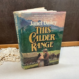 May include: A hardcover book titled "This Calder Range" by Janet Dailey. The cover features a landscape with a covered wagon and figures, with the title in large, gold lettering. The book is part of the Calder Saga series.