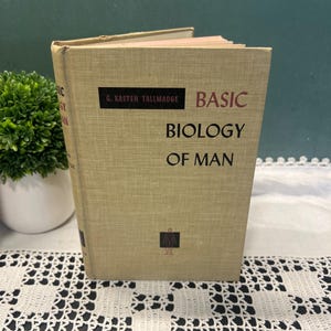 May include: A hardcover book titled Basic Biology of Man by G. Kasten Tallmadge with a tan, textured cover. The front cover features black text, a black rectangular accent, and a small black icon of a human figure centered at the bottom.