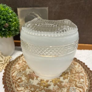 May include: A white glass light fixture with a decorative, textured top and a patterned band around the middle. The fixture sits on a patterned, round mat. A small green plant in a white pot is in the background.