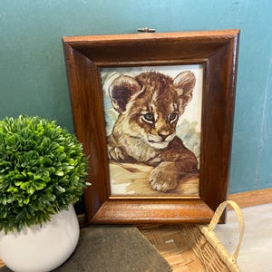 May include: A framed print of a lion cub. The artwork features a detailed watercolor-style illustration of a young lion with brown and tan fur. The frame is made of dark wood. A small green plant in a white pot is in the foreground.