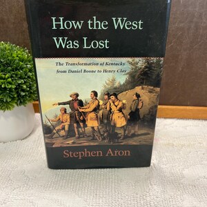 May include: A hardcover book titled "How the West Was Lost" with a black cover. The book's cover features an illustration of several men in period clothing. The author's name, Stephen Aron, is printed at the bottom.
