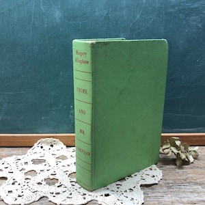 May include: A vintage green paperback book titled "Crime and Mr. Campion" by Margery Allingham. The book is sitting on a white doily on a wooden surface.