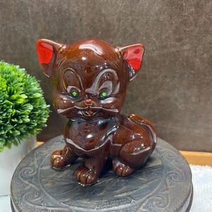 May include: A brown ceramic kitten figurine with red-tipped ears and green eyes. The vintage-style cat is in a seated position, with a glossy finish. The figurine is on a decorative, dark-colored surface.