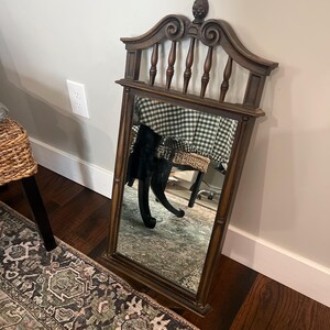 May include: A rectangular wall mirror with a dark brown wooden frame. The top section features a decorative arched pediment with vertical spindles and a central finial. The mirror glass is clear and reflects the surrounding room environment.
