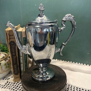 May include: A polished silver-toned metal coffee pot featuring an ornate handle, a decorative finial on the lid, and a tapered spout. The body of the pot is engraved with the text "NAOMI MILLER 1952" and stands on a circular pedestal base.
