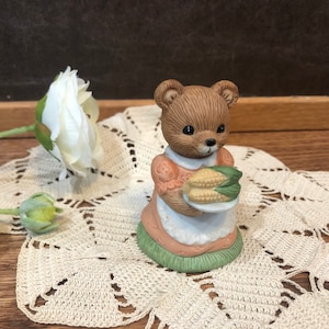 May include: A ceramic figurine of a brown bear wearing a pink dress and holding two ears of corn. The bear is standing on a green base.