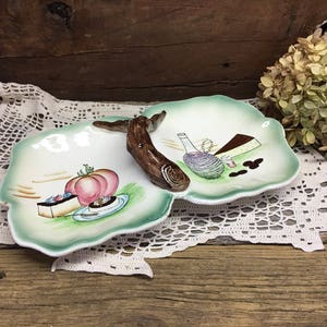 May include: A two-section serving dish with a green and white painted design. The dish features a painted wooden handle and images of fruit, pastries, and wine.