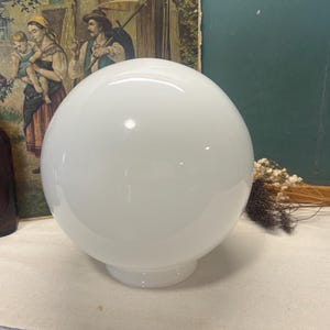 May include: A large, white, spherical glass lamp shade with a rounded base. The shade is opaque and reflects light. The background includes a vintage painting and a brown bottle.