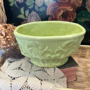 May include: A light green ceramic bowl with a pedestal base and embossed floral design. The bowl is sitting on a white lace doily and a small book. The background includes a wicker basket and floral artwork.
