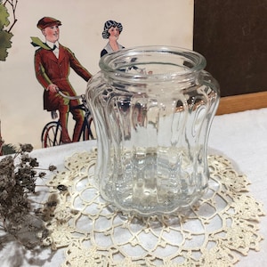 May include: A clear glass jar with a wide, ribbed body and a rounded base. The jar sits on a cream-coloured, lace doily. An antique illustration of a man on a bicycle is in the background.