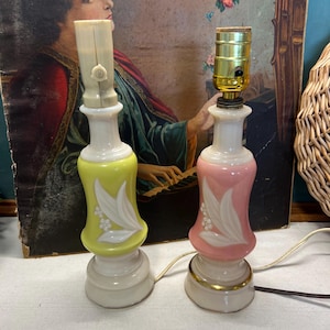 May include: Two vintage ceramic table lamps. One is yellow, the other is pink, both with white floral designs. The lamps have a white base and a cream-colored top. The pink lamp has a gold band around the base.