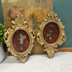 May include: Two ornate, gold-colored oval picture frames. Each frame has a red interior and a detailed, scrollwork design. The frames are displayed against a vintage background with a green surface and a white lace tablecloth.